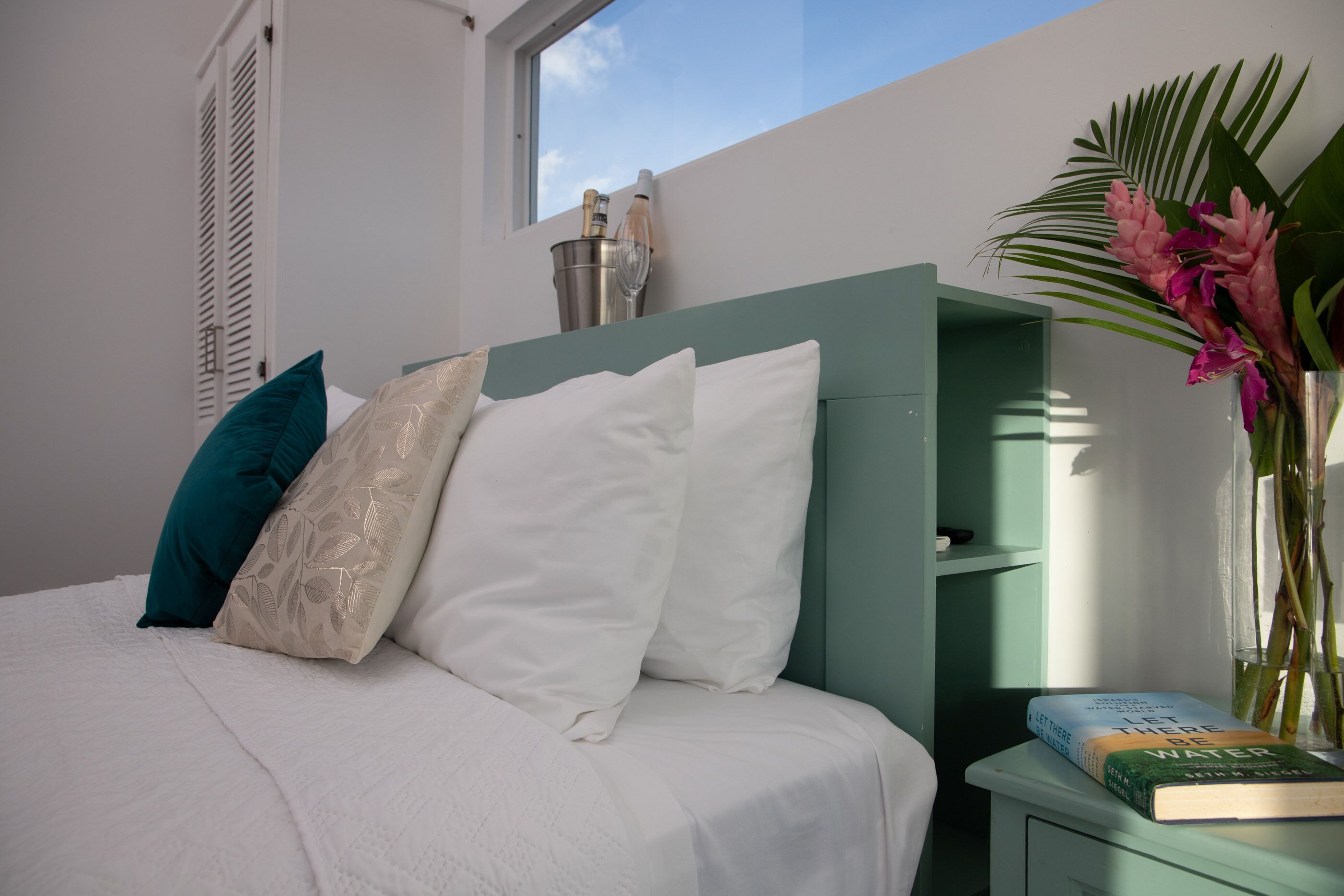 Una cama bien tendida con almohadas blancas y turquesa, una mesita de noche con un libro y un florero, y una ventana con vista al cielo.