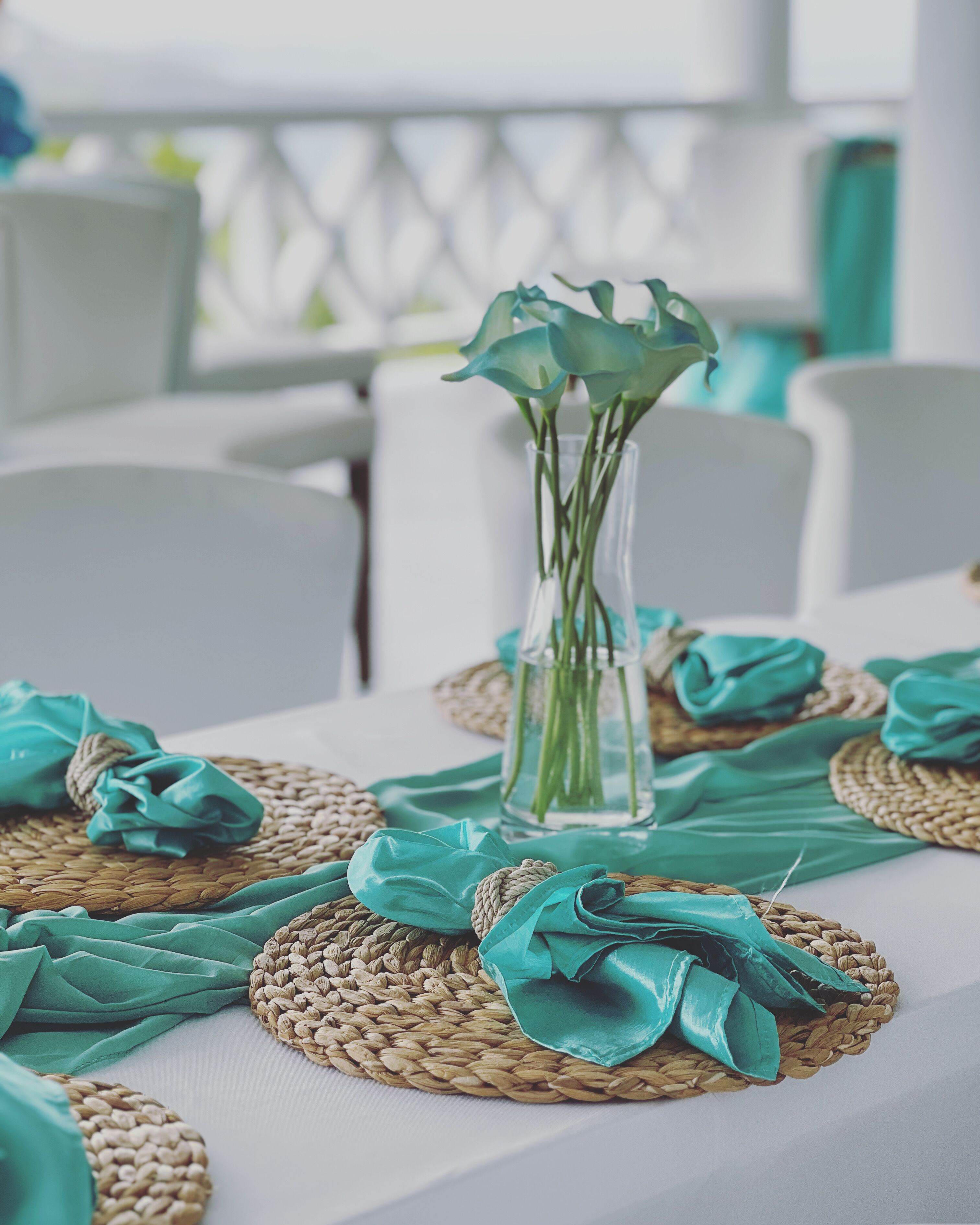 Una mesa vestida con individuales tejidos, un florero transparente con flores y servilletas color turquesa.