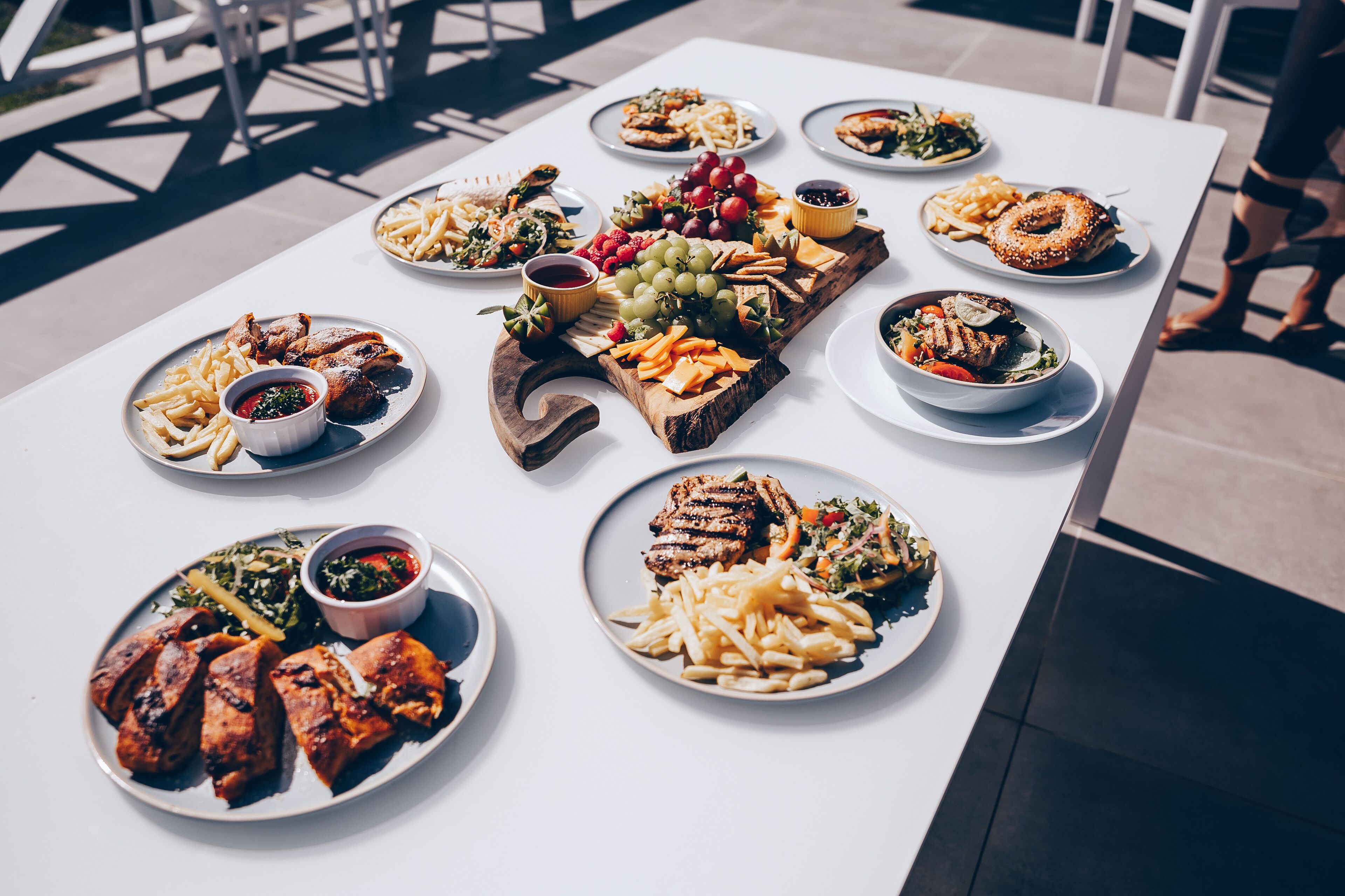 Een tafel met verschillende gerechten, waaronder vlees, friet en salades, buiten op een witte tafel.
