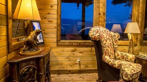 Interior acogedor de una cabaña con paredes de madera, un ventanal grande, una silla y una mesa con una lámpara.