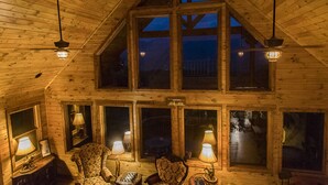 Interior de una cabaña de troncos con techos altos, ventanas amplias y varias lámparas.
