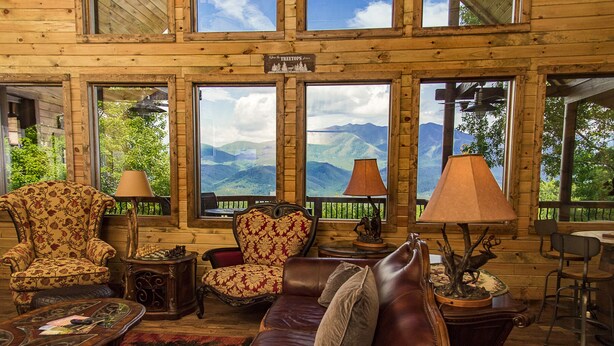Interior acogedor de una cabaña con sofá de cuero, sillón floral y vistas a las montañas a través de amplios ventanales.