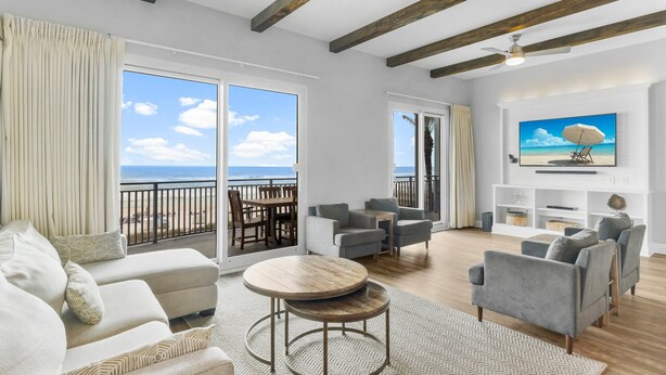 A spacious living room with a large window overlooking the beach, a round wooden table, and a flat-screen TV mounted on the wall. - Sterling Breeze 203 (Panama City Beach)