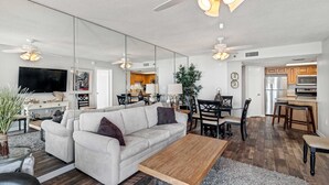 Una sala de estar moderna con un sofá, una mesa de centro y un comedor. Se aprecia una cocina al fondo con armarios de madera y un refrigerador.