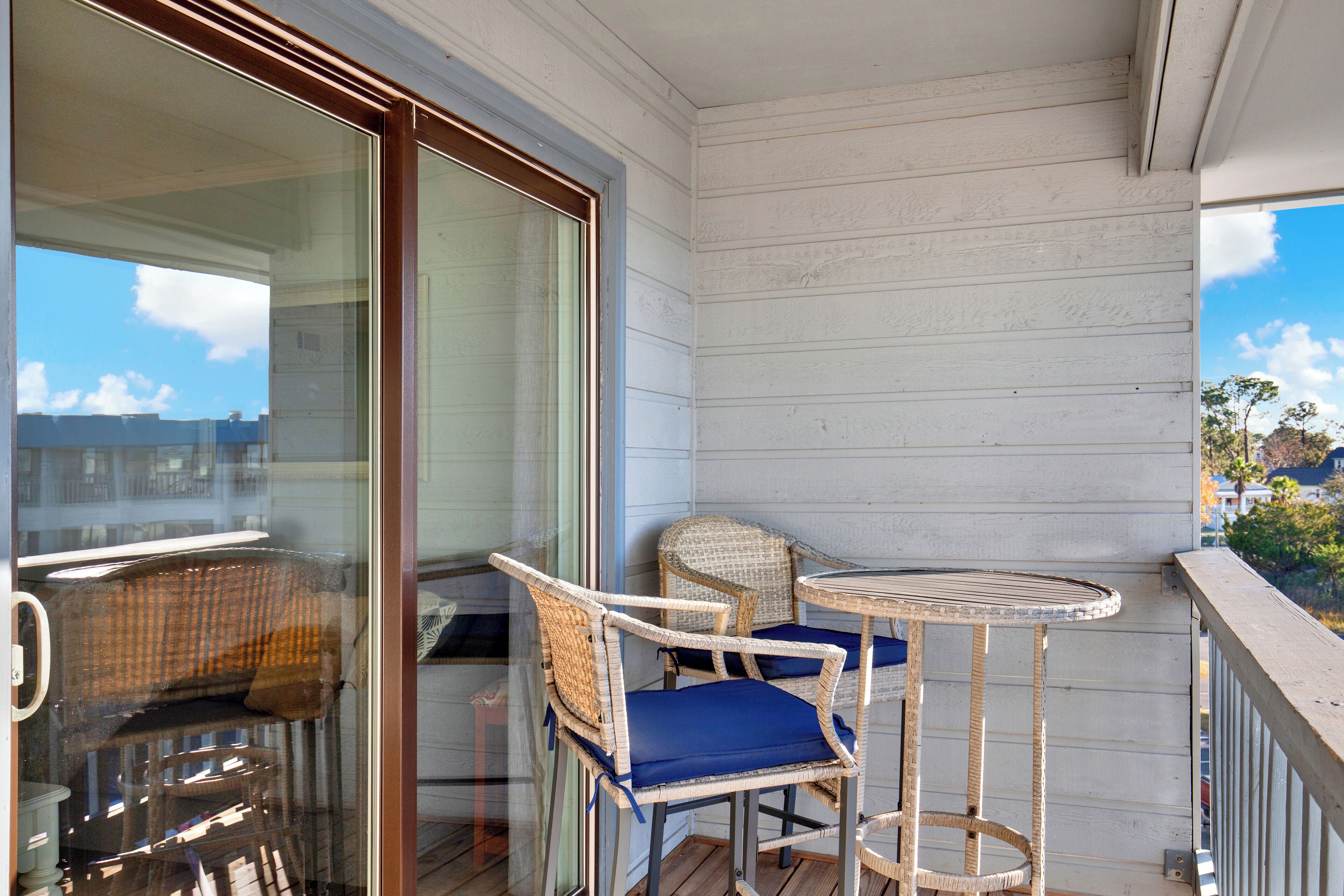 A balcony with wicker chairs and a small table, a sliding glass door, and a view of the sky and buildings.