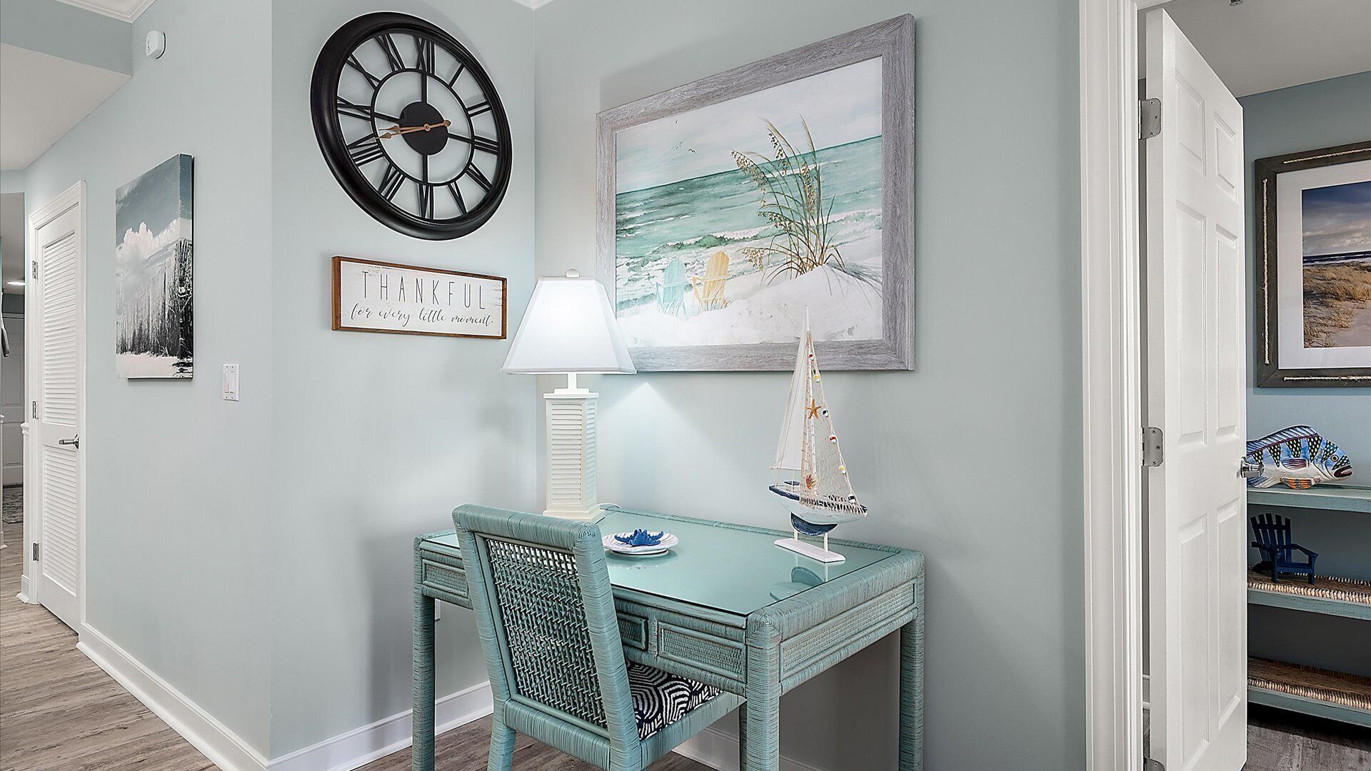 Un couloir avec un mur de couleur turquoise, une horloge murale, un tableau encadré, une petite table avec une lampe et une chaise.