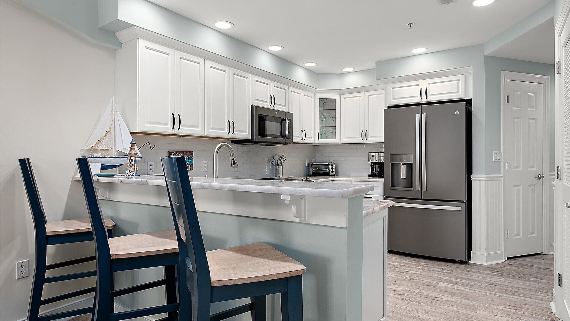 A modern kitchen with white cabinetry, a stainless steel refrigerator, a microwave, and a sink. There are two blue chairs with wooden seats.