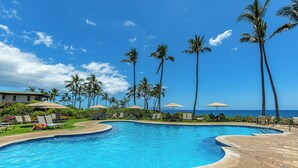 A pool area with lounge chairs and umbrellas, surrounded by palm trees and a view of the ocean.