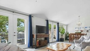 Un ampli saló amb un sofà, una taula de centre i una televisió sobre un moble de fusta. Hi ha grans finestres amb cortines, una zona de dinar amb cadires i un penjador.