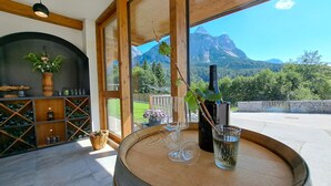 A wooden barrel table with wine glasses and a bottle, a potted plant, and a view of mountains through large windows. - New Apart at the Zugspitze with spa, hot tub, barbecue area, beautiful outdoor area... (Ehrwald)