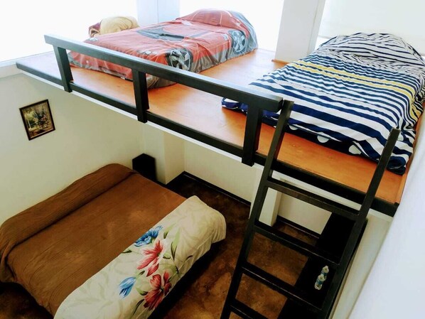 A dormitory-style room with two bunk beds, a wooden ladder, and a small seating area with a floral cushion.