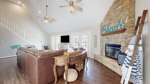 A spacious living room with a stone fireplace, a wooden dining table, a brown leather couch, and a staircase with white railings. - Beautiful Crystal Beach Get Away - Close to everything. 3 BR - Sleeps 10 (Bolivar Peninsula)
