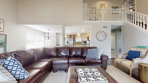 A spacious living room with a large brown leather sectional sofa, a coffee table with game pieces, and a dining area with a chandelier. - Beautiful Crystal Beach Get Away - Close to everything. 3 BR - Sleeps 10 (Bolivar Peninsula)