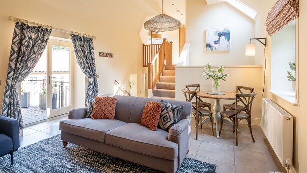 A cozy living room with a sofa, patterned curtains, a dining table, and a staircase leading to an upper level. - Sandsend - Rambling Rose -  a family break that sleeps 4 guests  in 2 bedrooms (Whitby)