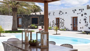 An outdoor pool area with a wooden table and chairs, surrounded by a white building with stone accents and a palm tree. - Hortensia, La Casa del Medianero (Macher)