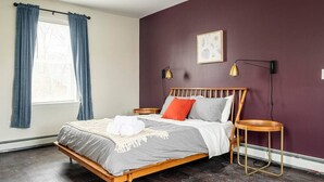 Een slaapkamer met een houten bed, een nachtkastje, een klein rond tafeltje, een raam met gordijnen en een wandlamp.