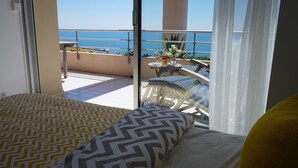 Un balcón con vista al mar, una mesa redonda con sillas y una cama con ropa de cama estampada.
