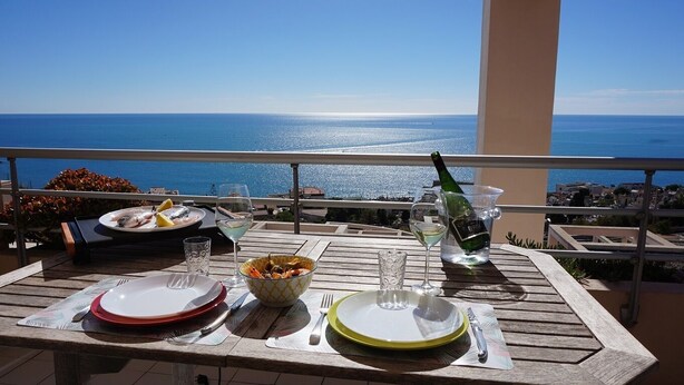 Una mesa con platos, cubiertos y una botella de champán en un balcón con vista al mar.