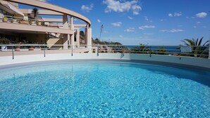 Una piscina azul clara con borde curvo, rodeada por un balcón con plantas en macetas y vista al océano.