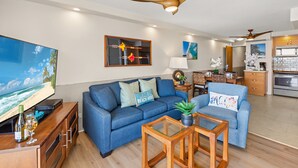 Uma sala de estar moderna com um sofá azul, uma mesa de centro de madeira e uma TV sobre um suporte de madeira. Há um ventilador de teto, uma área de cozinha com armários e uma mesa de jantar com cadeiras.