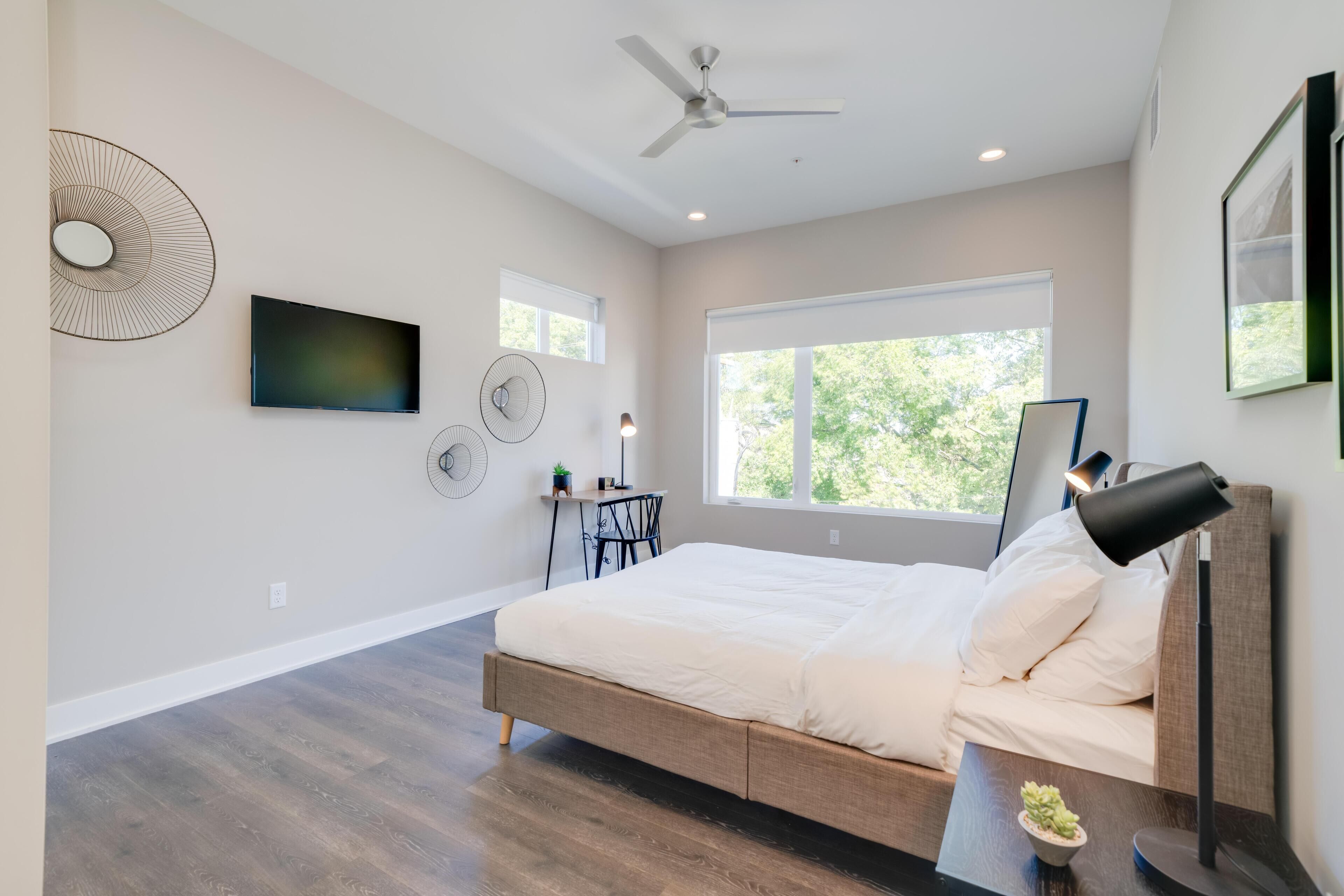 A modern bedroom with a bed, ceiling fan, wall-mounted TV, and large window.