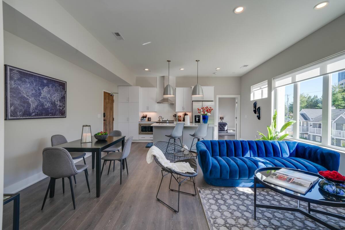 Ein modernes Wohnzimmer mit blauem Sofa, Essbereich mit Stühlen, einer Küche im Hintergrund und großen Fenstern.