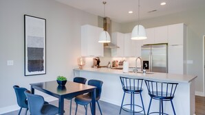 Una cocina moderna con armarios blancos, electrodomésticos de acero inoxidable e iluminación colgante. Hay un comedor con una mesa de madera oscura y sillas azules.