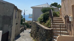 A narrow street with a stone wall and a staircase leading to a building with a balcony. - Maison au Centre de Pietranera. Premier Étage (San-Martino-di-Lota)