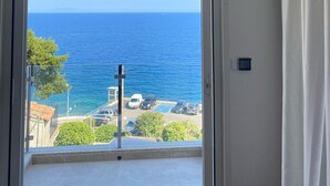 A room with a large sliding glass door leading to a balcony with a view of the sea and parked cars. - Maison au Centre de Pietranera. Premier Étage (San-Martino-di-Lota)