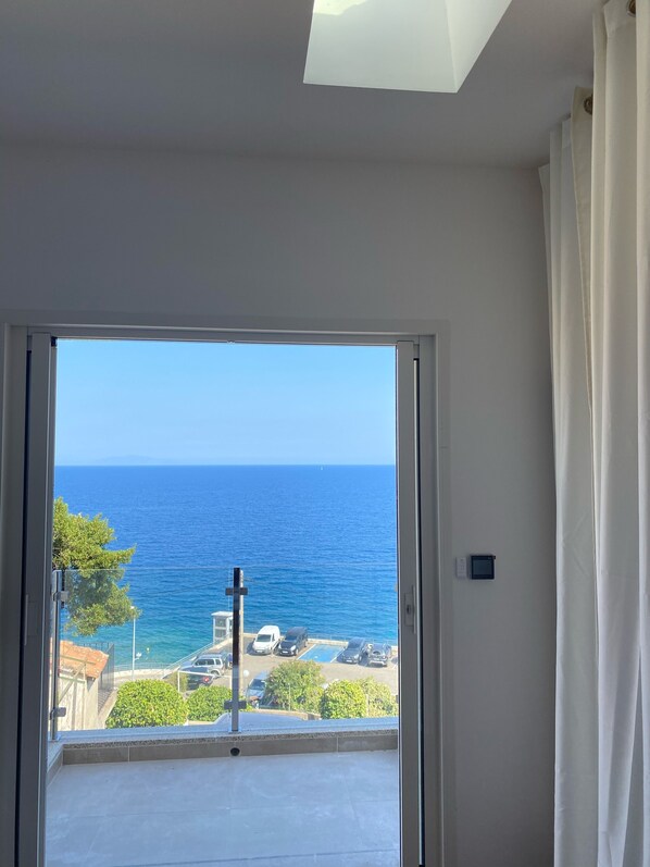 A room with a large sliding glass door leading to a balcony with a view of the sea and parked cars.