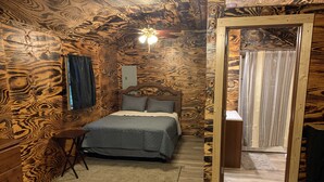 Un dormitorio con diseño de pared de madera, una cama con ropa de cama gris, una mesita peque ña y un ventilador de techo.