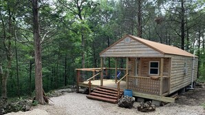 Una cabaña de troncos con terraza de madera y un pequeño porche, rodeada de un bosque.