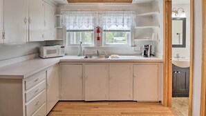 A compact kitchen with white cabinets, a microwave, a sink, and a window with lace curtains. - Homey Laconia Home w/ Private Porch & Grill (Laconia)