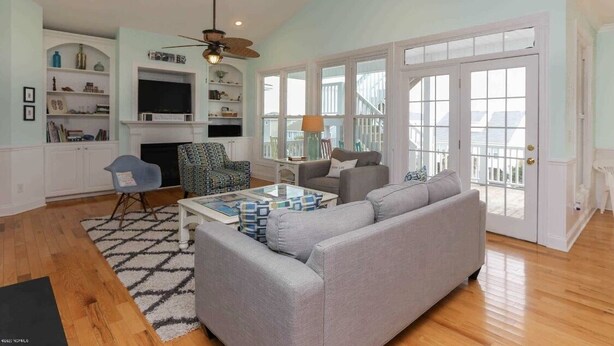 Un salón espacioso con un sofá gris, una mesa de centro y una chimenea. Hay estanterías con objetos decorativos y un televisor.