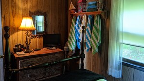 A wooden room with a bed, a television, a lamp, a shelf with towels, and a window with curtains. - Charming 2 Bedroom Lakefront Cottage in the Catskill Mountains (Roscoe)