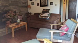 A cozy living room with a stone fireplace, a wooden coffee table, a brown sofa, and a blue armchair with a decorative pillow. - Charming 2 Bedroom Lakefront Cottage in the Catskill Mountains (Roscoe)