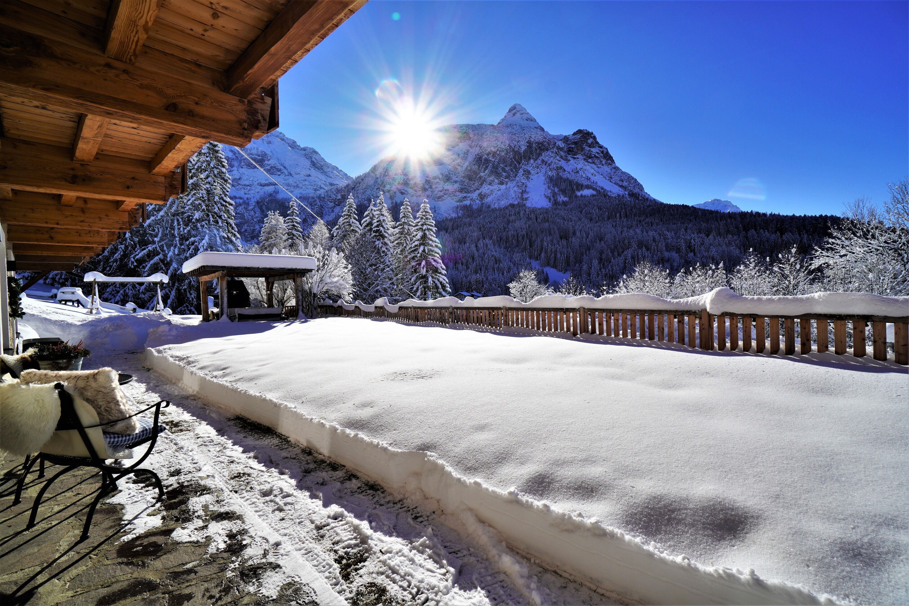A snow-covered outdoor area with a wooden deck, a covered seating area, and a mountain backdrop.