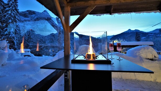A wooden structure with a glass table displaying a lit fire and two glasses of red wine.