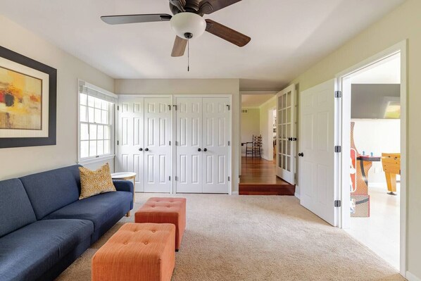 A living room with a dark blue sofa, two orange ottomans, a ceiling fan, and a large window. - Over 10 Acres In Bucolic Bucks County w/Pool! (Perkasie)
