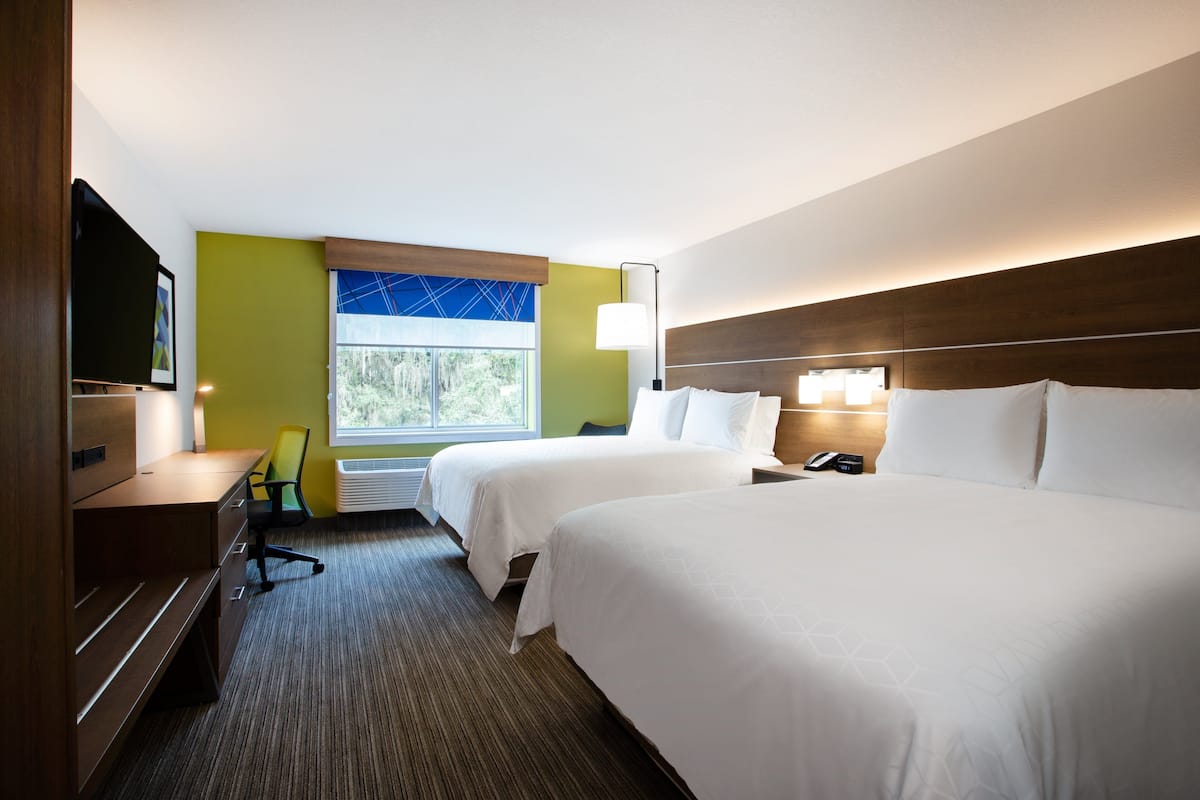 A hotel room with two beds, a desk, a chair, a TV, and a window with a view of trees.