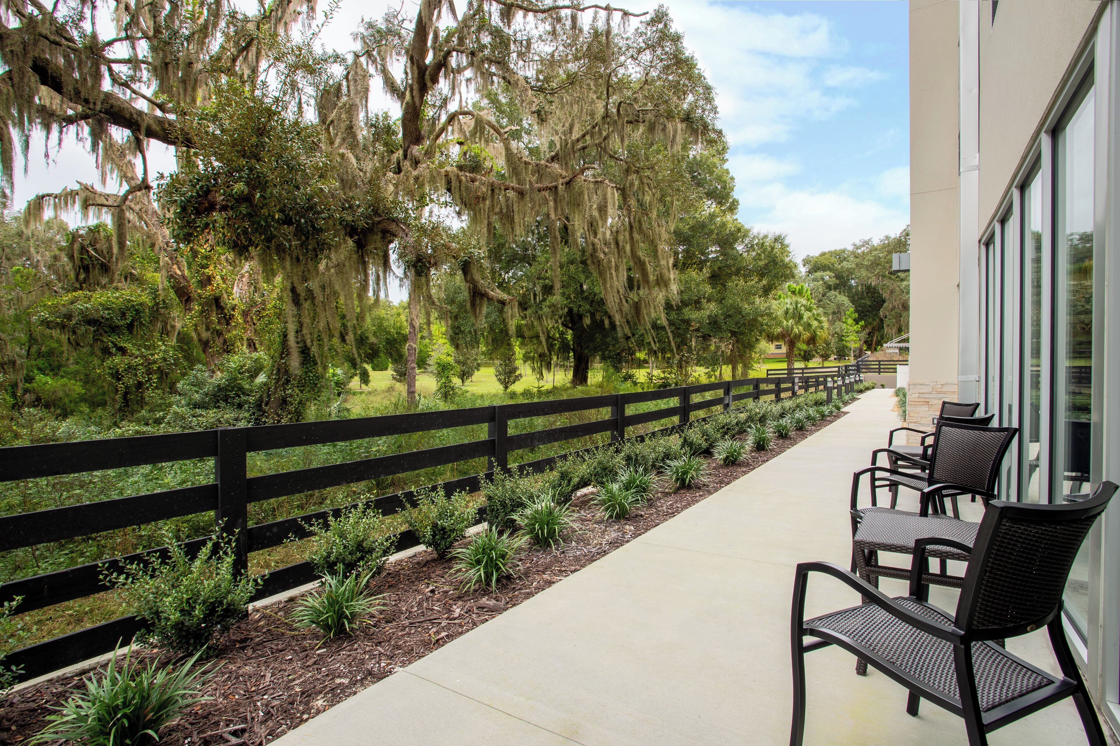 A covered outdoor seating area with a black fence, chairs, and a pathway lined with plants.
