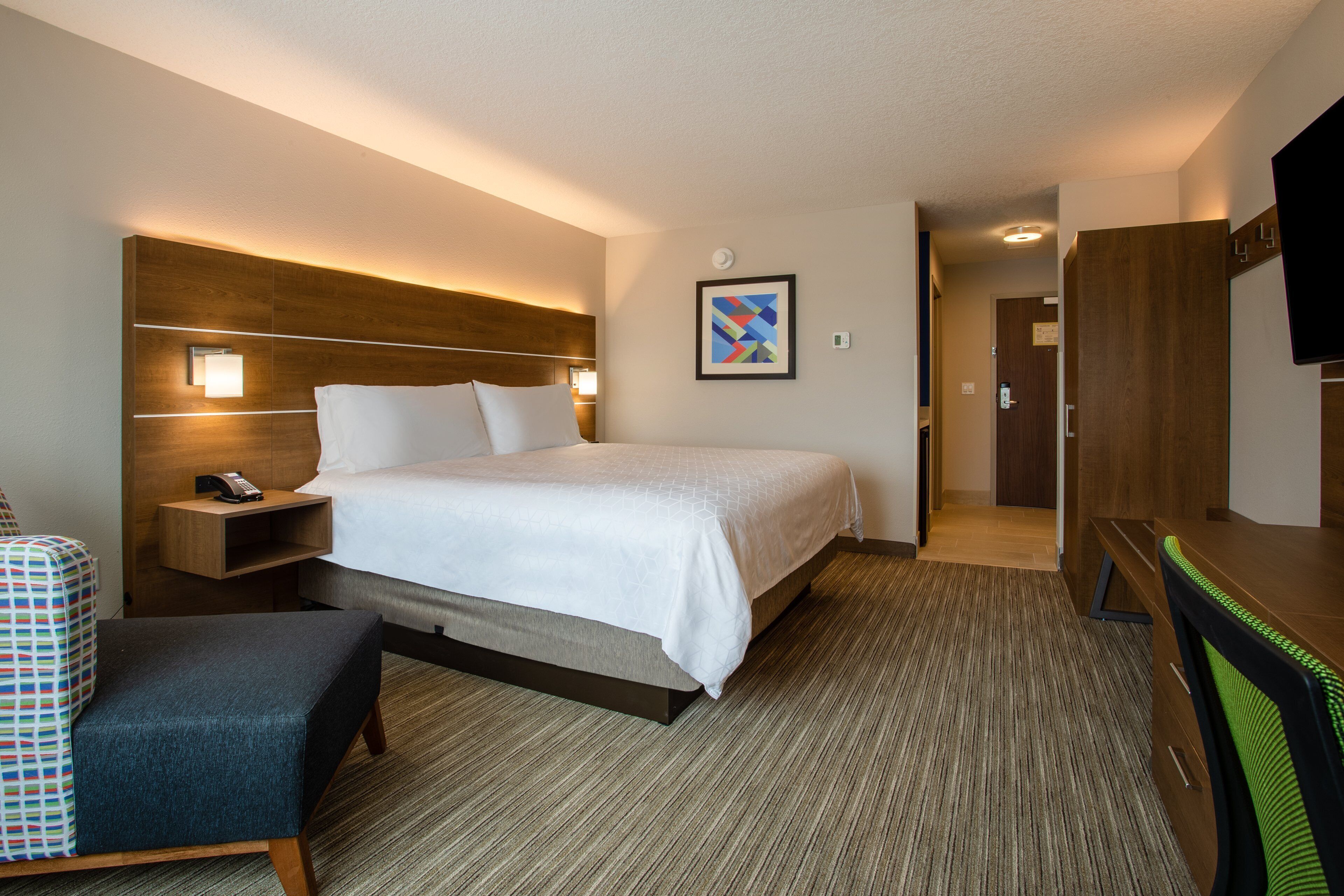 A hotel room with a large bed, a desk, a chair, a bedside table with a phone, and a TV on the wall.