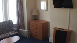 A hotel room with a sofa, a wooden dresser, a television mounted on the wall, and a window with curtains. - Glen Villa Flat 1 - 4 Berth (Great Yarmouth)