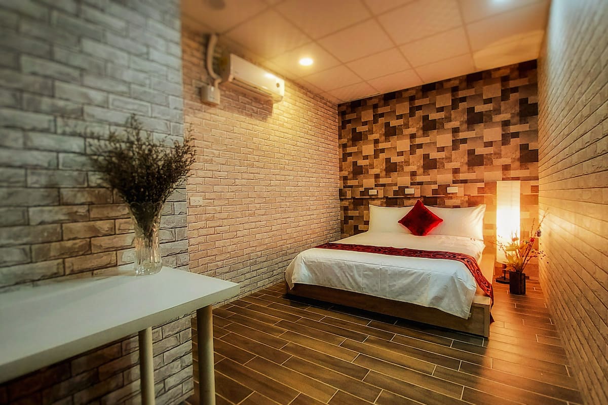 A hotel room with a brick wall, a wooden floor, a bed with white bedding and a red pillow, a small table with a vase, and a wall-mounted air conditioner.