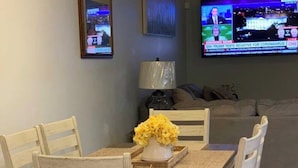 A dining area with a wooden table and chairs, a vase with yellow flowers, and a television displaying news. - Comfort Zone Palace Inn (Jacksonville)