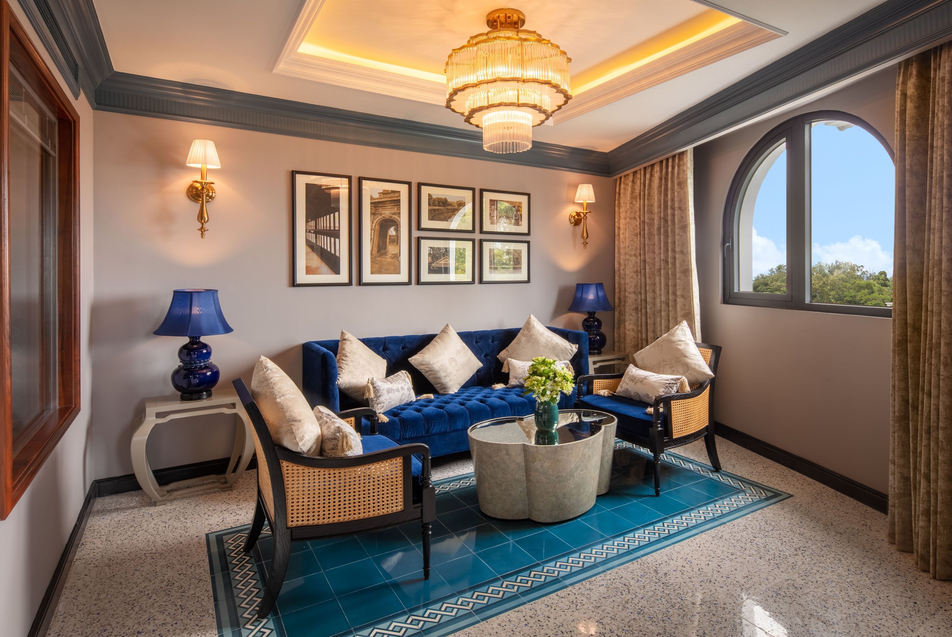 Un salon avec un canapé bleu, deux tables d’appoint avec des lampes, un lustre, des tableaux encadrés au mur et une grande fenêtre avec des rideaux.