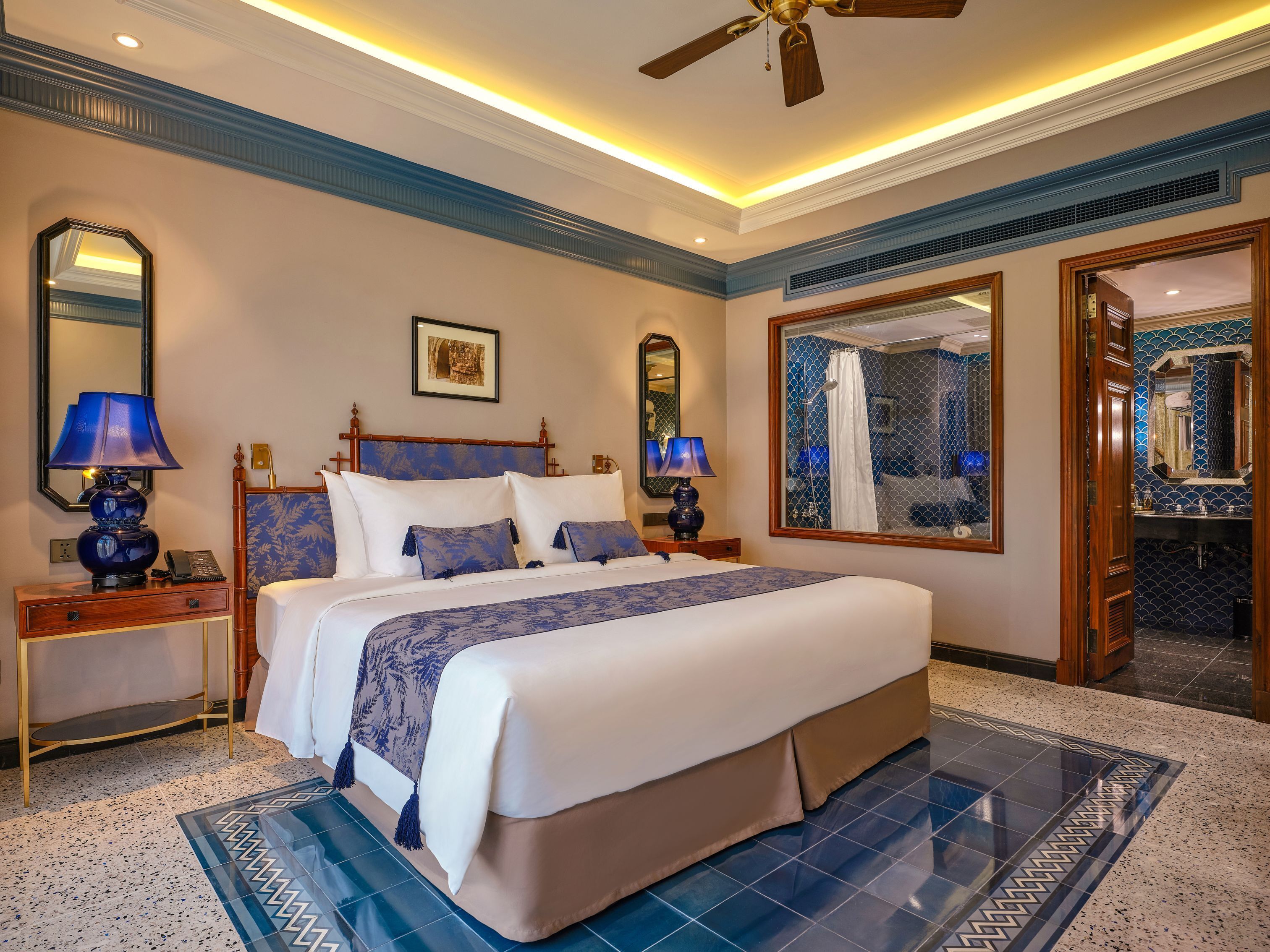 A hotel room with a large bed, two bedside tables with lamps, a ceiling fan, a large mirror, and a bathroom visible through a glass door.