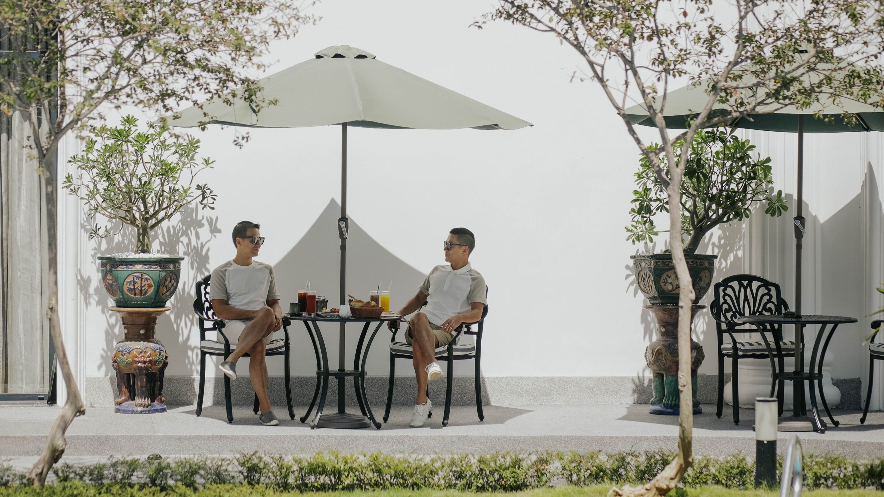 Two men seated at a table with a drink and food, under a large umbrella, with potted plants and a white wall in the background.