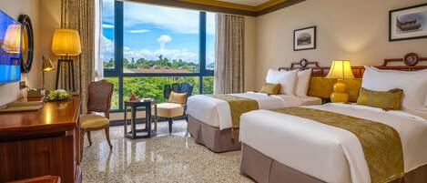 A hotel room with two beds, a desk, a chair, a TV, and a large window with a view of greenery and buildings.