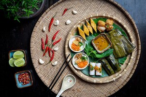 A traditional meal with various dishes on a woven mat, including rice cakes, dumplings, and a bowl of sauce.